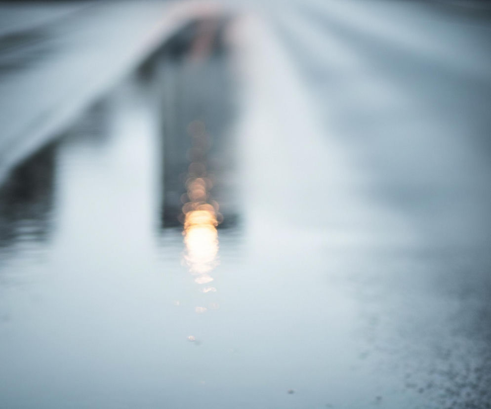 Rozmyty widok mokrej, asfaltowej powierzchni drogi, z perspektywy bliskiej ziemi. W centrum, na drodze, widoczne jest podłużne odbicie jasnego, pomarańczowo-żółtego światła, składające się z rozmazanych, okrągłych punktów. Powierzchnia drogi jest wilgotna, miejscami tworząc kałuże, które odbijają otoczenie w postaci ciemnych, nieostrych kształtów po lewej stronie kadru oraz pojedyncze, blade światła w tle. Ogólna kolorystyka jest chłodna, z dominującymi odcieniami szarości i błękitu, kontrastującymi z ciepłym blaskiem centralnego światła.