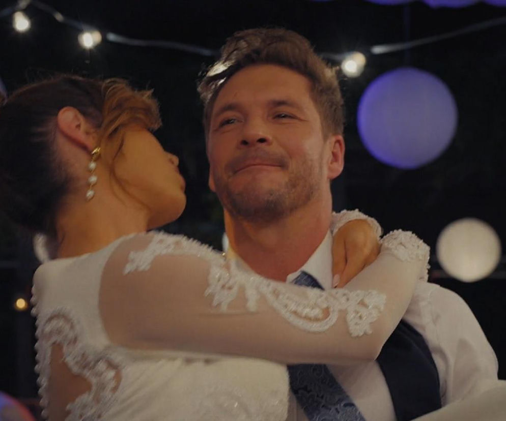 Marcin Chodakowski (Mikołaj Roznerski) trzyma na rękach Kamę (Michalina Sosna), oboje uśmiechnięci, w strojach ślubnych, w tle widoczne ozdobne lampki i lampiony na wieczornym przyjęciu weselnym. Fotografia ilustruje dramatyczne wydarzenia z M jak miłość, o których przeczytasz na portalu Super Seriale.