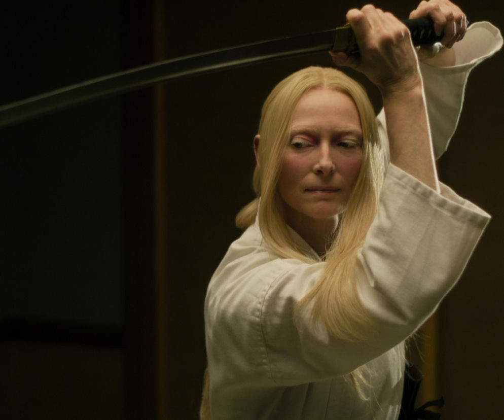 Aktorka Tilda Swinton w białym stroju trzyma miecz samurajski obiema dłońmi. O jej wystawie w Warszawie przeczytasz w Eska.