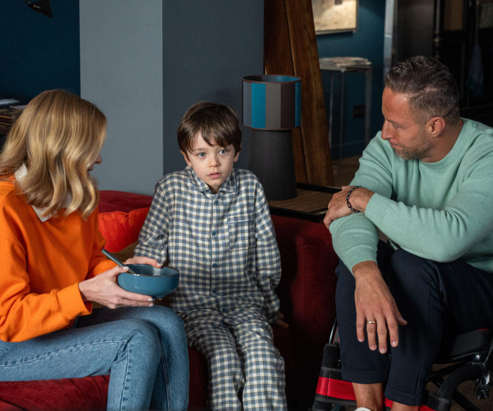 Dominika, Mateuszek i Sebastian z serialu „Barwy szczęścia” siedzą na czerwonej kanapie, dyskutując o trudnej sytuacji. Dominika trzyma miskę, Mateuszek w piżamie patrzy z niepokojem. Sebastian, siedzący na wózku inwalidzkim, patrzy na Mateuszka, jak przygotować go do zeznań. Więcej o ich losach na Super Seriale.