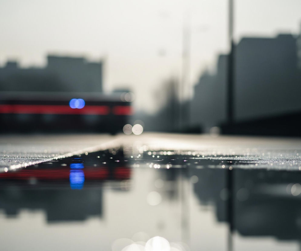 Ulica po deszczu, jej powierzchnia w dolnej połowie zdjęcia odbija niewyraźne światła i kształty. Na pierwszym planie widoczne są drobne kropelki wody i odblaski, tworzące punkty świetlne. W tle, rozmyty, czerwony pojazd z dwoma wyraźnymi, niebieskimi światłami przednimi, porusza się po mokrej nawierzchni. Po obu stronach ulicy zarysowują się ciemne bryły budynków, również rozmyte.