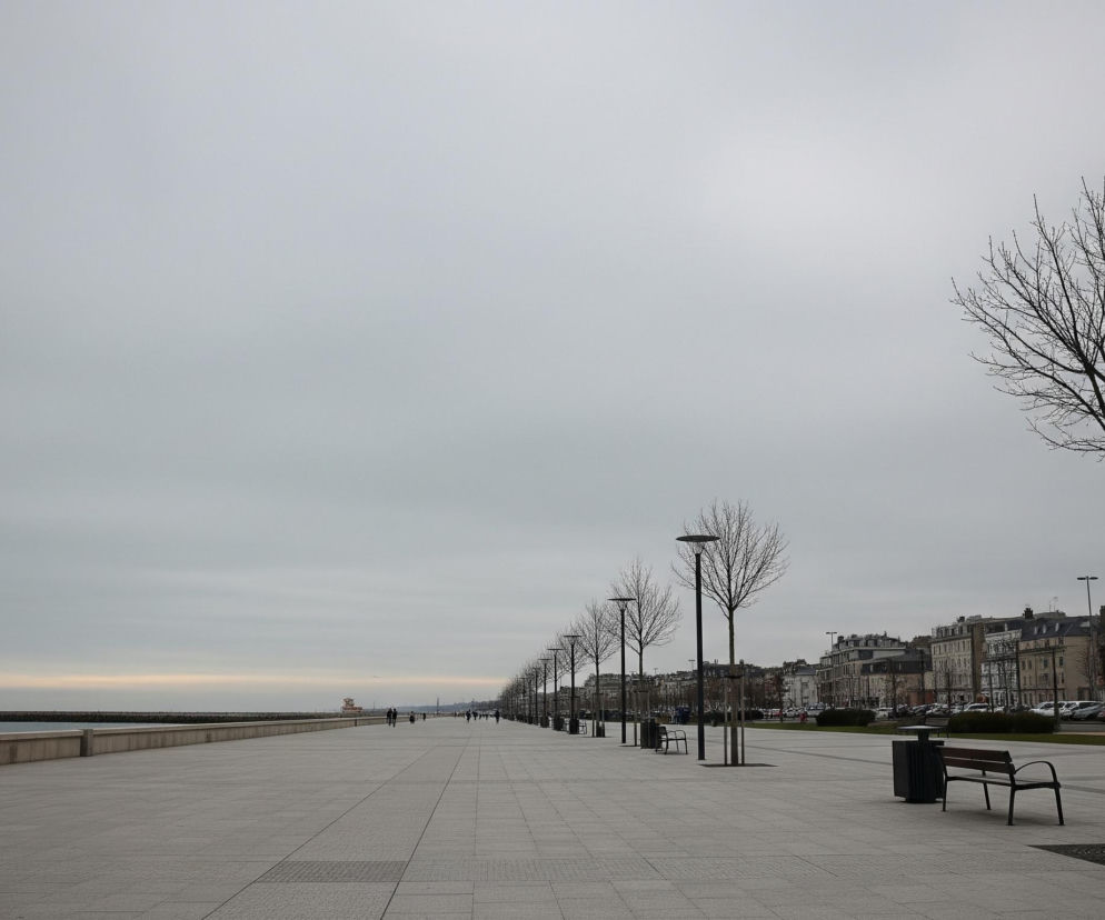 Długa, brukowana promenada biegnie wzdłuż prawego do lewego dolnego rogu zdjęcia, z widocznymi rzędami łysych drzew i latarni, które wznoszą się po prawej stronie. Po lewej stronie promenady rozciąga się morze z falochronem i małą latarnią morską w oddali, natomiast po prawej stronie widoczne są budynki miejskie, wzdłuż których zaparkowane są samochody. Niebo jest pochmurne i szare, jedynie u dołu horyzontu lekko rozświetlone jasnym, beżowym pasmem.