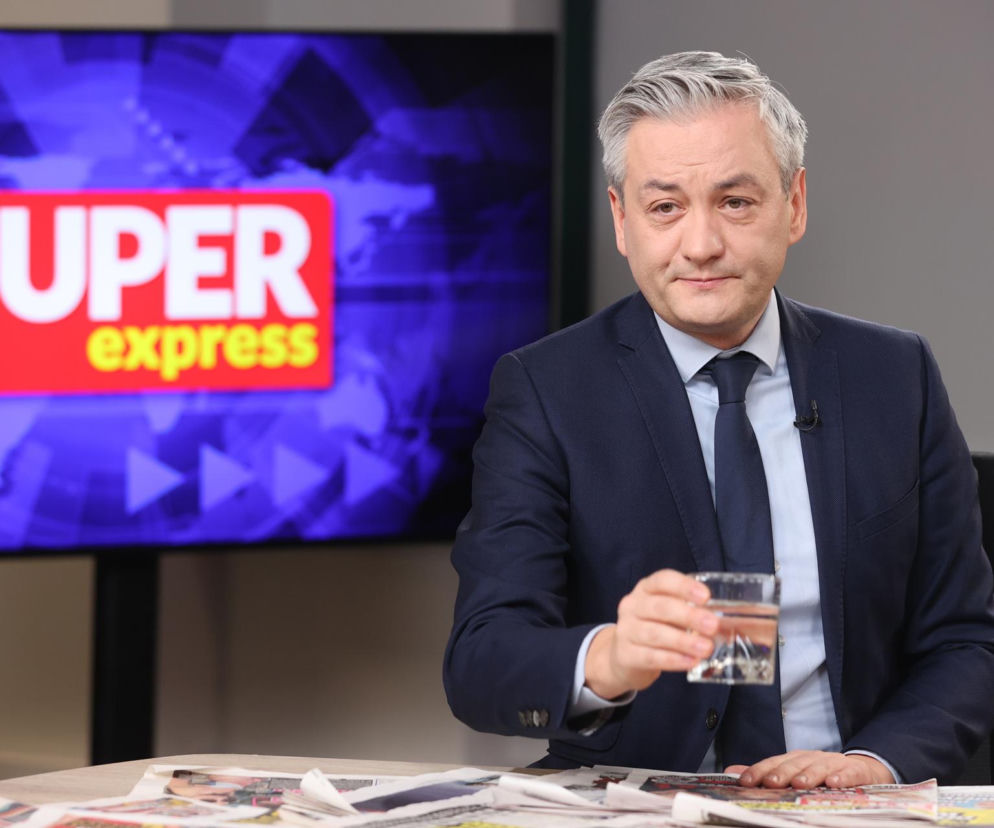 Robert Biedroń w granatowym garniturze i krawacie, z siwymi włosami, siedzi za stołem, trzymając szklankę wody. W tle ekran z logo Super Express. Polityk krytykuje politykę rządu i porusza temat relacji polsko-ukraińskich, o czym można przeczytać na naszym portalu.