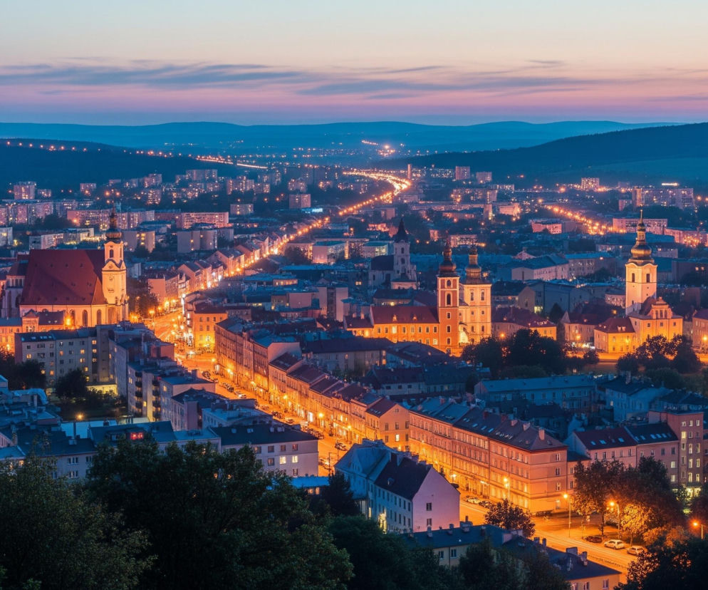 Rozległa panorama miasta o zmierzchu, widziana z góry, z licznymi rozświetlonymi budynkami i ulicami, biegnącymi w kierunku oddalonych, ciemnych wzgórz. Dominują ciepłe, pomarańczowe światła lamp ulicznych i oświetlonych okien, kontrastujące z granatowo-fioletowym niebem i ciemnozielonymi drzewami na pierwszym planie. W centrum kadru wznoszą się wieże kościelne i czerwony dach dużej budowli, a w tle widać autostradę wijącą się przez miasto, oświetloną rzędem świateł.