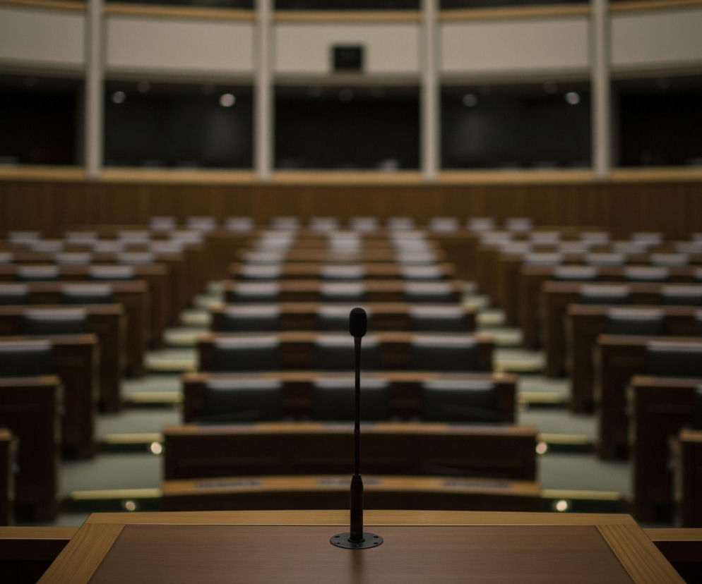 Na pierwszym planie widoczny jest brązowy drewniany pulpit z ciemniejszym blatem, na którym stoi czarny, długi mikrofon na elastycznym ramieniu, zakończony okrągłą główką. Za pulpitem rozciąga się pusta, amfiteatralna sala wykładowa lub konferencyjna, z rzędami drewnianych ław i ciemnych krzeseł, skierowanych w stronę pulpitu. Ściany są wykończone panelami w kolorze drewna, a u góry sali znajduje się rząd ciemnych okien lub wnęk, przedzielonych jasnymi, pionowymi elementami. Oświetlenie w sali jest stonowane, dominują ciemne brązy i szarości, z kilkoma jasnymi punktami w tle.