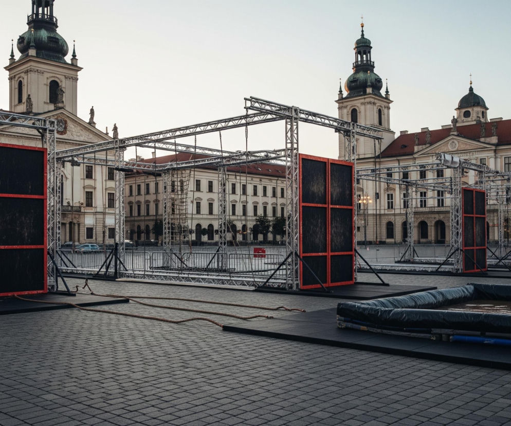Na pierwszym planie widoczny jest brukowany plac, na którym ustawiono konstrukcje ze stalowych kratownic. Po lewej stronie znajdują się dwie duże, prostokątne tablice z czarnego materiału, obramowane czerwonymi liniami, zawieszone na metalowych ramach. Pomiędzy nimi i za nimi rozciągają się kolejne elementy tych kratownicowych konstrukcji, tworząc rodzaj toru przeszkód.

Po prawej stronie, na czarnych matach, leżą długie, czarne wały, a za nimi widać płytkie baseny z wodą, również obramowane czarnym materiałem. W tle dominują majestatyczne budynki z jasnymi elewacjami i czerwonymi dachami, a nad nimi wznoszą się dwie wieże kościelne z zielonymi kopułami i złotymi krzyżami, wszystko pod jasnym, bezchmurnym niebem.