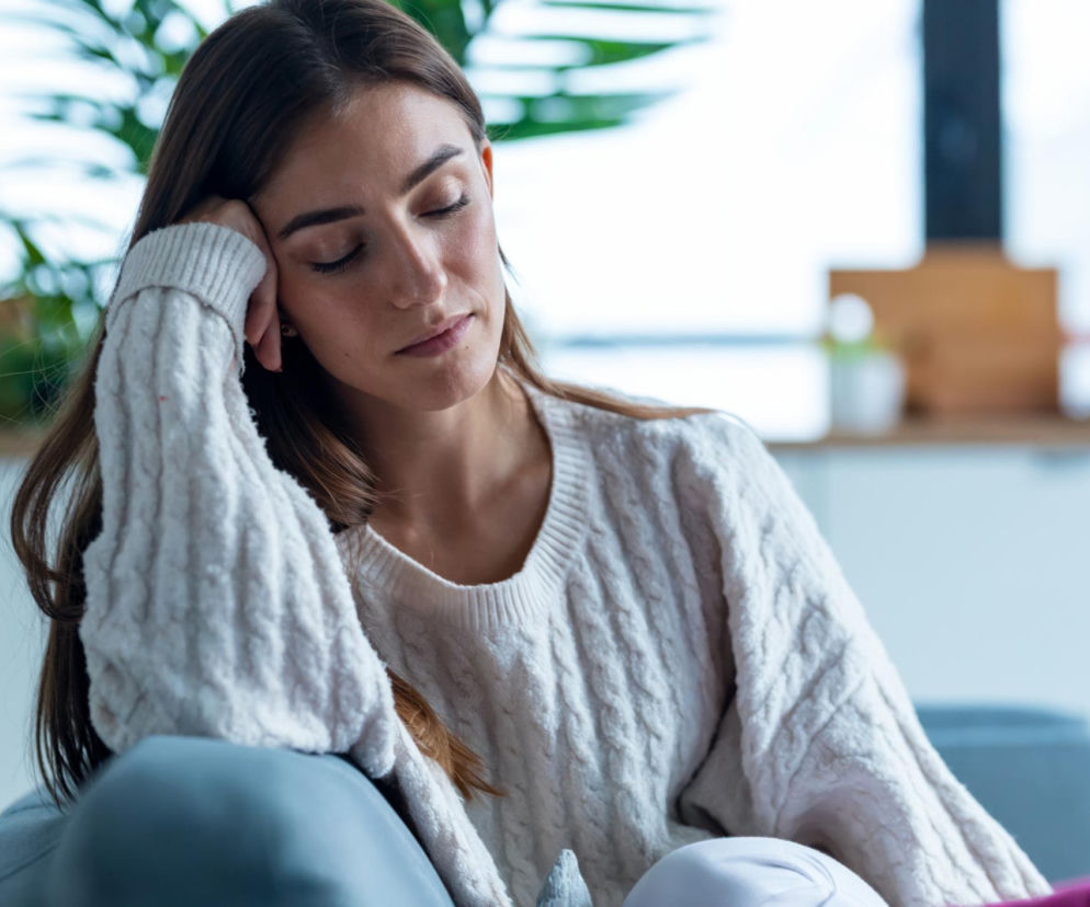 Młoda kobieta z długimi, ciemnymi włosami, w jasnym, wełnianym swetrze, z zamkniętymi oczami, opiera głowę na dłoni, siedząc na kanapie. Wygląda na zmęczoną, zatroskaną lub pogrążoną w refleksji, co sugeruje stany depresyjne lub przewlekły stan zapalny, o którym możesz przeczytać na Poradnik Zdrowie.