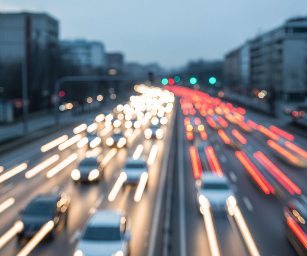 Długa ekspozycja uchwyciła ruch samochodów na wielopasmowej autostradzie w nocy lub o zmierzchu. Po lewej stronie jezdni widoczne są smugi białego światła reflektorów, a po prawej stronie smugi czerwonego światła tylnych lamp. Samochody są rozmazane, jednak widoczne są ich zarysy w jasnych, srebrzystych i ciemnych kolorach. W tle po obu stronach drogi majaczą niewyraźne, szare budynki oraz drzewa, a na niebie widoczne są zamglone chmury.