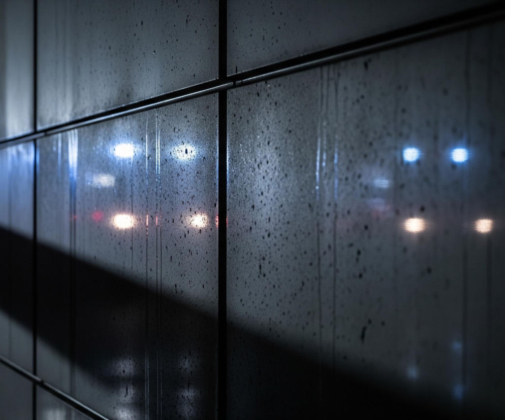 Ciemną ścianę pokrytą drobnymi kroplami, przypominającą szybę lub metalową powierzchnię. Widoczne są poziome i pionowe linie podziału, dzielące ścianę na prostokątne panele. W tle, za ścianą, odbijają się rozmazane, jasne światła: cztery biało-niebieskie punkty oraz cztery pomarańczowo-czerwone, sugerujące ruch drogowy. Ciemny cień w kształcie ukośnego pasa, tworzący trójkąt, rozciąga się od lewej górnej części obrazu w dół do środka.