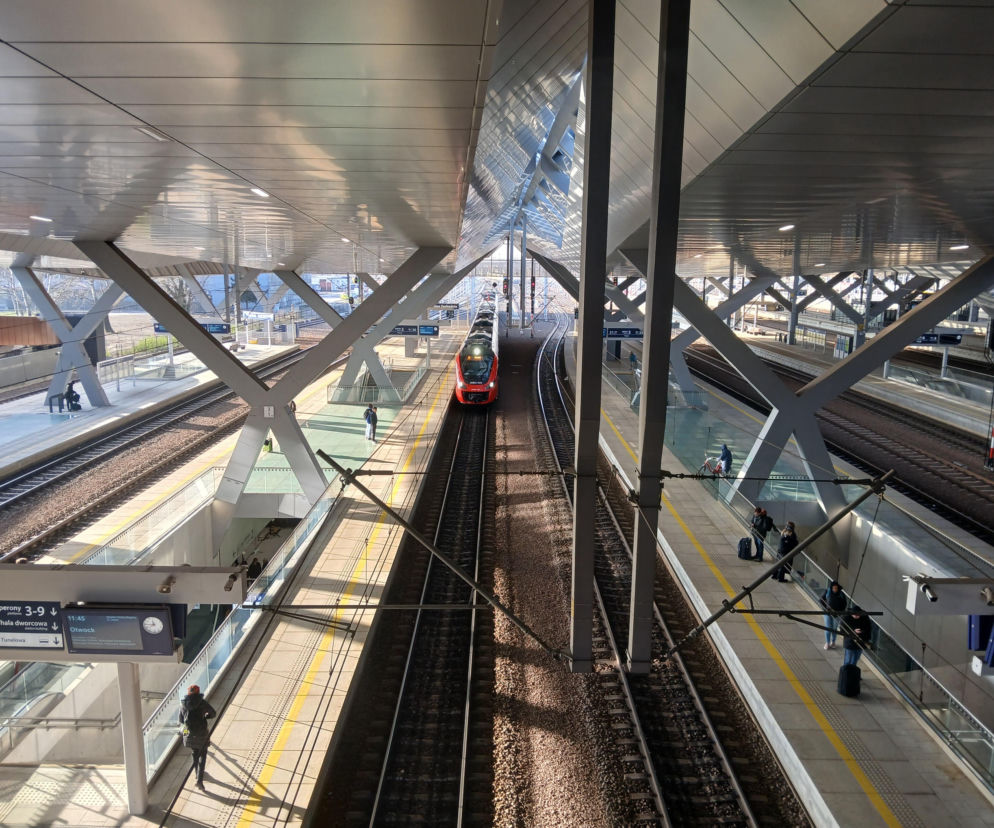 Zmodernizowany Dworzec Zachodni w Warszawie, z widocznymi peronami i torami kolejowymi, gdzie perony są zadaszone nowoczesnym, zintegrowanym dachem z funkcjonalnymi świetlikami. Pasażerowie czekający na pociągi, w oddali widoczny jest nadjeżdżający czerwony pociąg. Więcej o architekturze węzła kolejowego na Architektura Murator Plus.