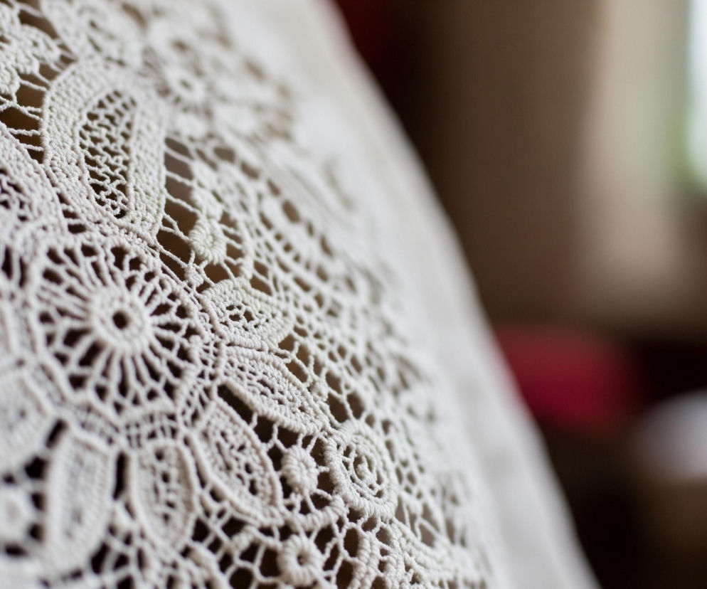 Na pierwszym planie, z lewej strony, widoczna jest biała, koronkowa tkanina o skomplikowanym wzorze, obejmująca większość kadru. Materiał ma gęsty, ażurowy splot, tworzący symetryczne, kwiatowe i geometryczne motywy z licznymi drobnymi oczkami. Prawa strona zdjęcia jest rozmyta, ukazując niewyraźne tło składające się z odcieni brązu, bieli i jasnej zieleni.