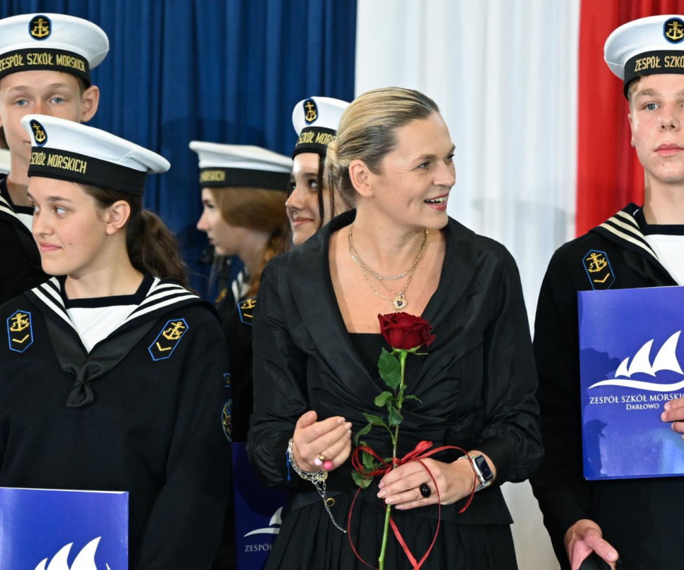 Barbara Nowacka, Minister Edukacji, w czarnym komplecie z naszyjnikami i pierścionkiem, uśmiechnięta, z czerwoną różą, stoi wśród uczniów Zespołu Szkół Morskich w Darłowie ubranych w granatowe mundury marynarskie. O wizycie minister przeczytasz na naszym portalu.