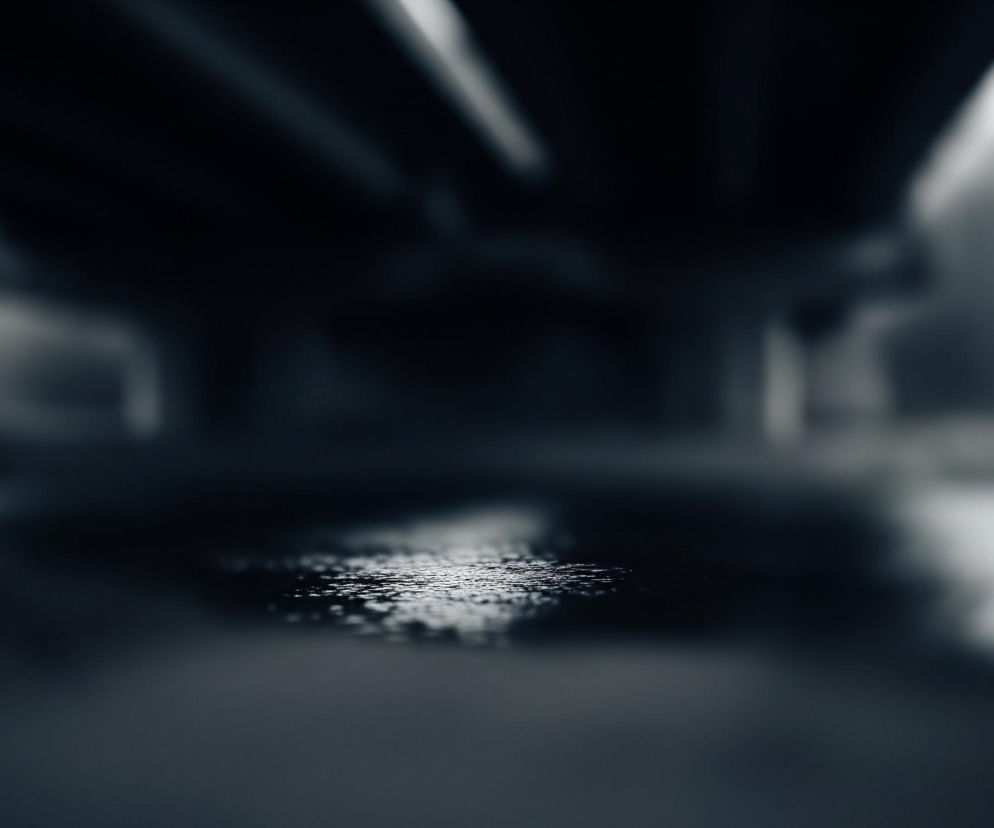 W centrum obrazu znajduje się kałuża wody, z widocznymi refleksami światła tworzącymi błyszczące punkty na jej powierzchni. Kałuża jest otoczona ciemnym, rozmytym gruntem, a dalej w tle widać ciemne, niewyraźne kształty przypominające konstrukcję mostu lub wiaduktu. Dominują ciemne odcienie szarości i granatu, z jaśniejszymi, rozmytymi elementami w górnej części kadru oraz na dalekim tle, sugerującymi mglisty lub pochmurny dzień. Całość kompozycji cechuje niewielka głębia ostrości, z ostrym punktem na kałuży i silnym rozmyciem tła.