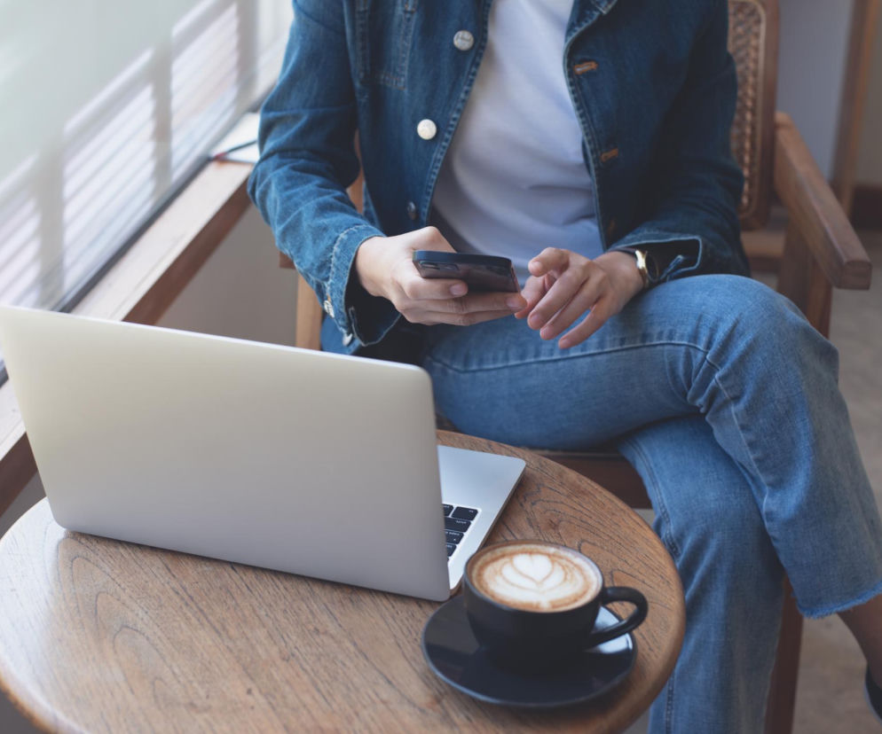 Kobieta w jeansowej kurtce i spodniach, siedząca na drewnianym krześle z założoną nogą na nogę, trzymająca smartfon. Na drewnianym stoliku przed nią znajduje się laptop i filiżanka kawy latte. Ta pozycja może prowadzić do przeciążeń i bólu pleców, o czym przeczytasz na Poradnik Zdrowie.