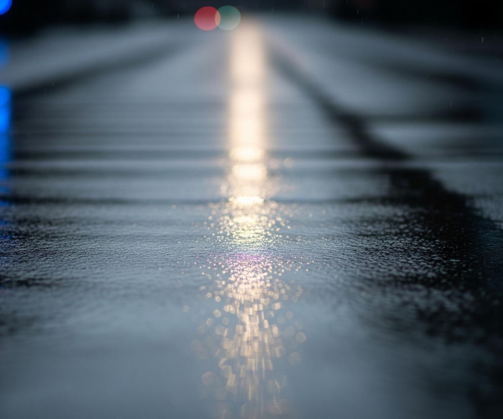 Mokry asfalt ulicy rozciąga się w głąb obrazu, pokryty licznymi refleksami świateł odbijającymi się od kałuż i mokrej nawierzchni. Pośrodku jezdni biegnie jasna, rozmyta linia światła, przechodząca od złotego w dolnej części do jaśniejszego, kremowego w oddali. Po lewej stronie, w tle, widoczne są intensywne, rozmyte światła w kolorach czerwonym i niebieskim, odbijające się również na mokrej powierzchni drogi. W górnej części obrazu, bardziej centralnie, znajdują się dwa rozmyte okrągłe światła: jedno czerwone i jedno zielone. Cały obraz utrzymany jest w ciemnych, chłodnych barwach z dominacją odcieni szarości, błękitu i czerni, przełamanych jaskrawymi plamami światła.