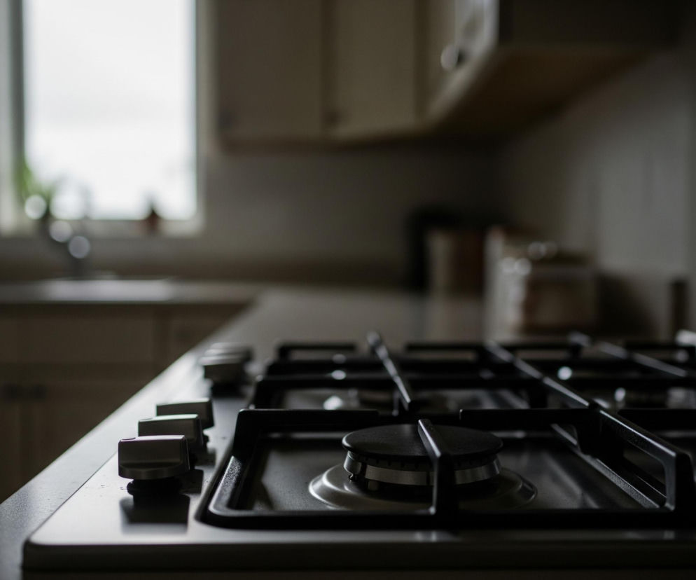 Na pierwszym planie, z prawego dolnego rogu, znajduje się stalowa płyta gazowa z czterema czarnymi palnikami i czterema pokrętłami do regulacji płomienia. Powierzchnia płyty odbija światło, tworząc refleksy. Tło jest rozmazane, przedstawiając wnętrze kuchni z jasnym oknem po lewej stronie i kilkoma górnymi szafkami w kremowym odcieniu. Cały obraz utrzymany jest w stonowanych, ciemnych barwach, z wyjątkiem jasnego, nieostrego okna.