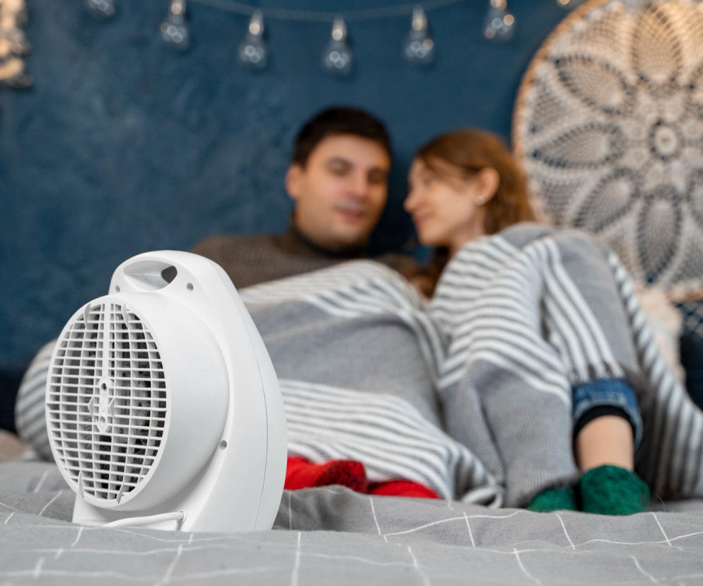 Biały wentylator elektryczny stoi na szarym łóżku z pościelą w kratę, a w tle widać rozmazaną parę pod kocem. To zdjęcie ilustruje, jak korzystać z wentylatora w sypialni, o czym przeczytasz więcej na Poradnik Zdrowie.