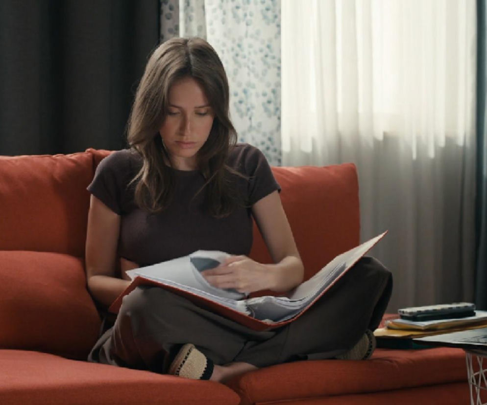 Młoda kobieta, Madzia (Natalia Sierzputowska) z serialu Barwy szczęścia, siedzi na pomarańczowej sofie, przeglądając dokumenty. Na jej twarzy widać zamyślenie i troskę, co odzwierciedla jej trudną sytuację finansową. Więcej o losach Madzi na portalu Super Seriale.