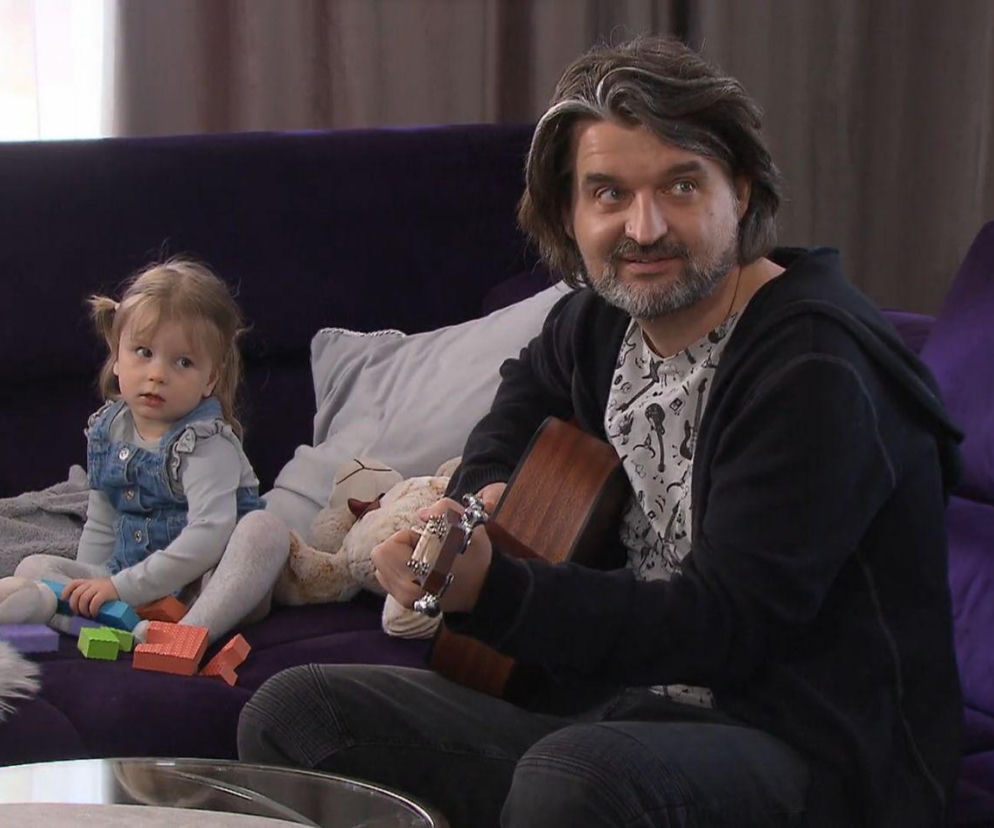 Mężczyzna z brodą i siwymi pasemkami we włosach, ubrany w ciemną bluzę i koszulkę z nadrukiem, gra na gitarze siedząc na fioletowej kanapie. Obok niego mała dziewczynka w dżinsowej sukience bawi się klockami. Zdjęcie z serialu Klan, a więcej o tym przeczytasz na Super Seriale.