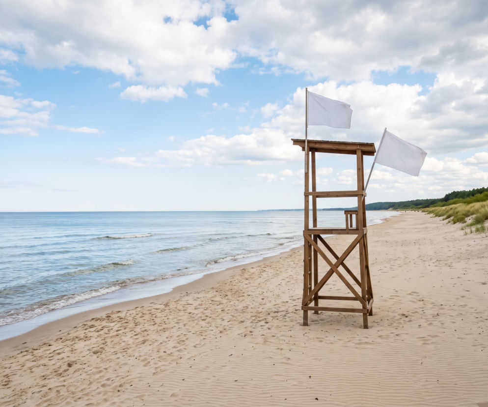 Drewniana wieża ratownicza z dwiema białymi flagami na pustej plaży. O pogodzie w Ustroniu Morskim przeczytasz na SE Szczecin.