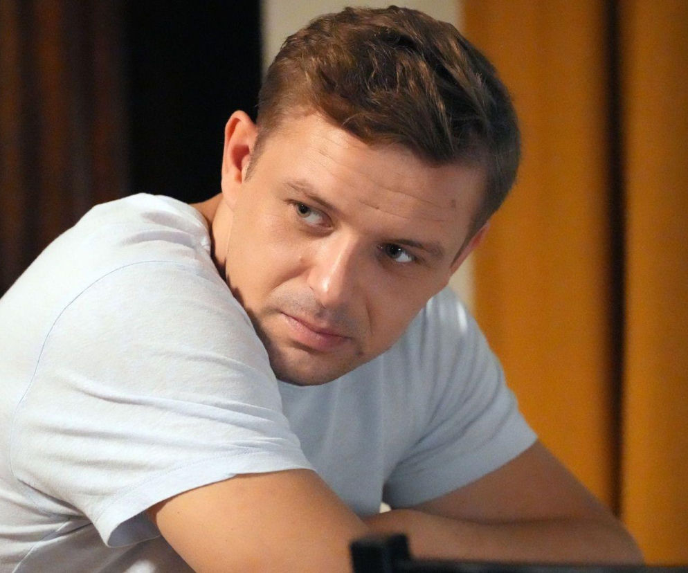 Marek Molak jako Hubert Pyrka w jasnym T-shircie, z zamyślonym wyrazem twarzy, odwracający głowę, co oddaje jego zaniepokojenie i stres związany z kłopotami finansowymi, o których można przeczytać na Super Seriale. W tle widać ciemne i pomarańczowe zasłony.