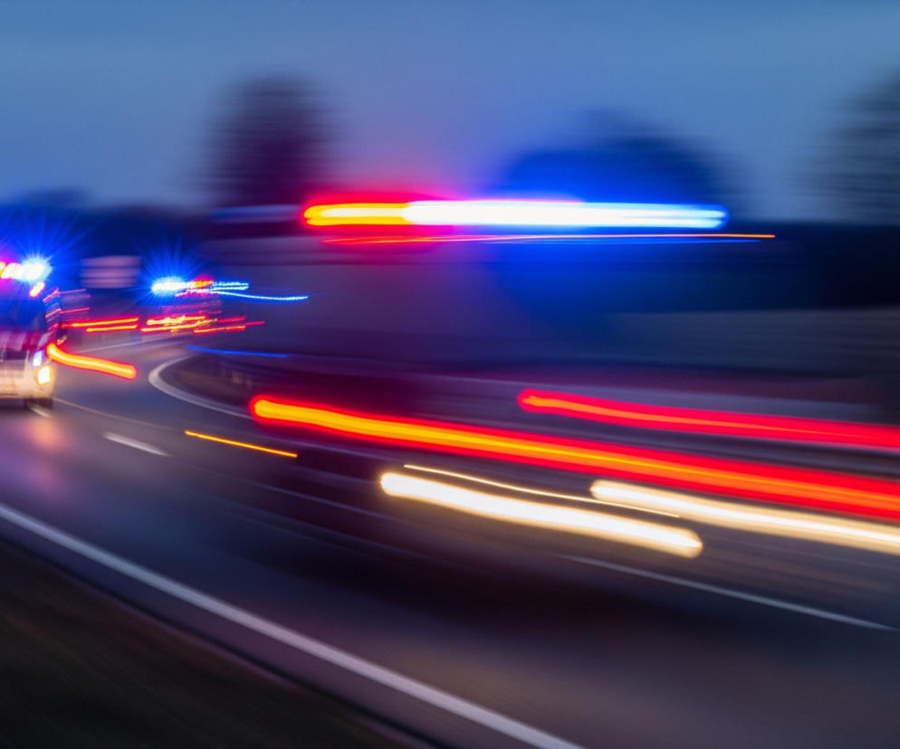 Biały pojazd uprzywilejowany z migającymi niebieskimi światłami awaryjnymi porusza się po asfaltowej drodze w lewo, z rozmyciem wskazującym na dużą prędkość. Za nim, po tej samej drodze, podąża drugi, mniej widoczny pojazd z migającymi czerwonymi i niebieskimi światłami. Po prawej stronie, na równoległej drodze lub pasie, widoczne są rozciągnięte smugi czerwonych i białych świateł, sugerujące ruch innych pojazdów w przeciwnym kierunku. Tło jest ciemne, niebo w odcieniach granatu, z niewyraźnymi ciemnymi konturami drzew po bokach drogi.