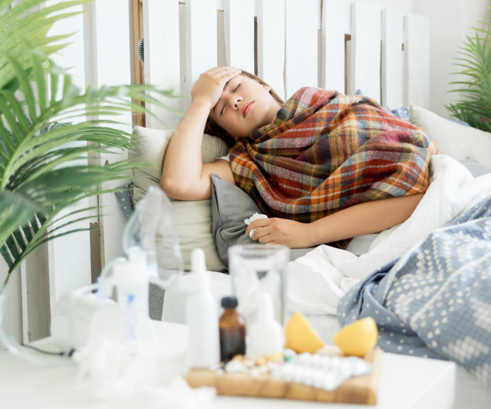 Młoda kobieta w kratkowanym kocu leży w łóżku z zamkniętymi oczami i dłonią na czole, sugerując chorobę, być może grypę wariantu K. Na stoliku nocnym widoczne są leki, szklanka wody i cytryna, co podkreśla tematykę walki z infekcją. Więcej o objawach grypy przeczytasz na Poradnik Zdrowie.