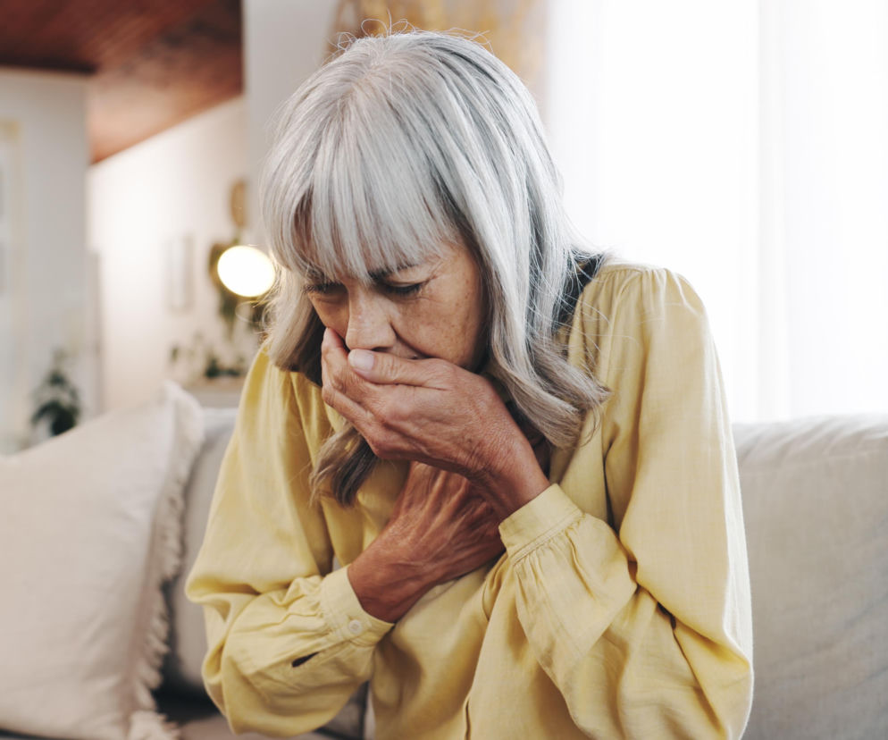 Starsza kobieta z siwymi włosami, ubrana w żółtą koszulę, kaszle, zakrywając usta dłonią. Druga dłoń spoczywa na jej klatce piersiowej, sygnalizując dolegliwości oddechowe. Zdjęcie ilustruje problemy z drogami oddechowymi, o których możesz przeczytać na portalu Poradnik Zdrowie.