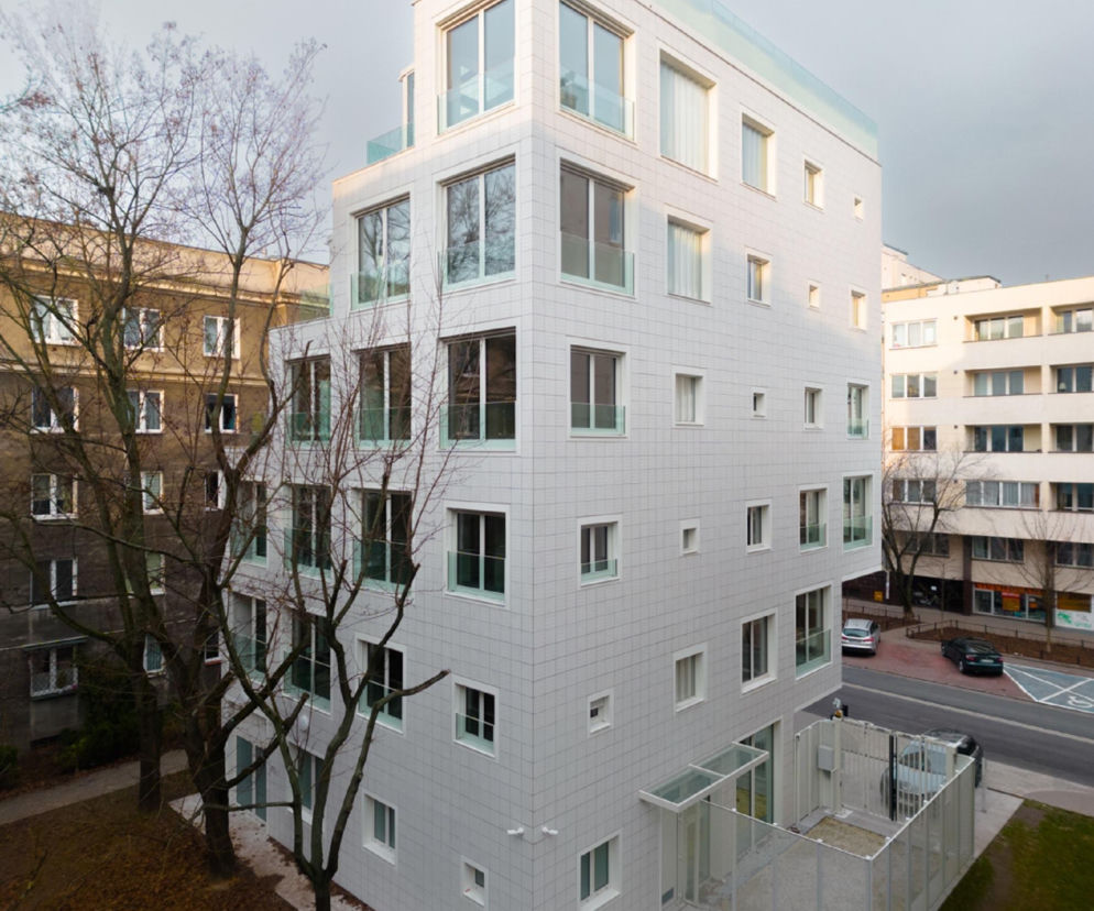 Nowoczesny budynek mieszkalny La Scala autorstwa POLE Architekci, z białymi ceramicznymi płytkami elewacyjnymi i dużymi oknami, wkomponowany w miejską zabudowę Warszawy. Szklane balustrady tarasów i dyskretna zieleń wokół obiektu podkreślają estetykę realizacji, o której możesz przeczytać na Architektura Murator Plus.