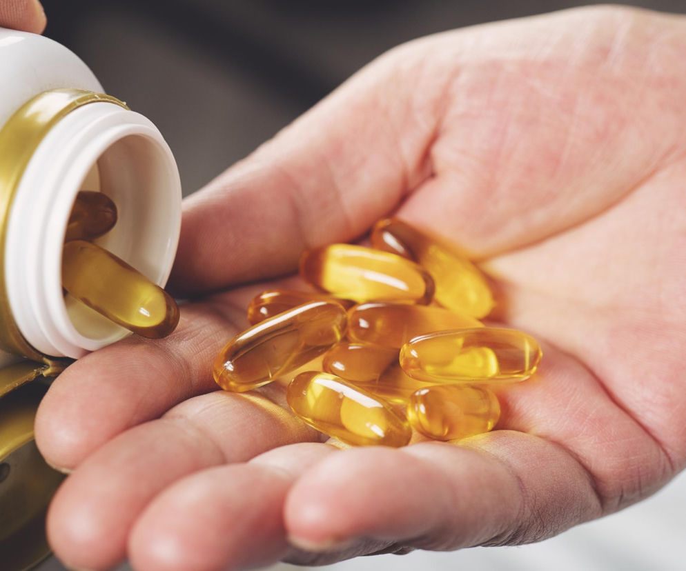 Żółte, owalne kapsułki witaminy D wysypujące się z białego pojemnika z złotym wieczkiem na otwartą dłoń. To nawiązanie do badania nad wpływem witaminy D na ograniczenie ryzyka long COVID, o czym przeczytasz więcej na Poradnik Zdrowie.