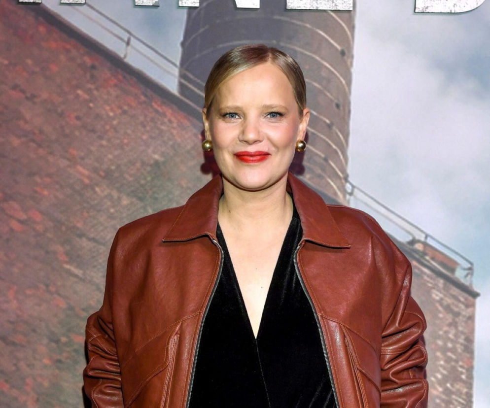 Joanna Kulig na zdjęciu portretowym, uśmiechnięta, w brązowej skórzanej kurtce i czarnym topie, z subtelnym makijażem i czerwonymi ustami. Jej włosy są gładko zaczesane. Aktorka pozuje na tle industrialnego budynku. Więcej o jej metamorfozie przeczytasz na naszym portalu.