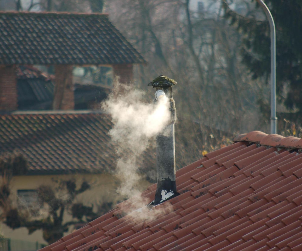 Gęsty dym wydobywa się z komina na dachu domu z czerwoną dachówką. O kontrolach pieców przeczytasz na Eska Ostrzeszów.