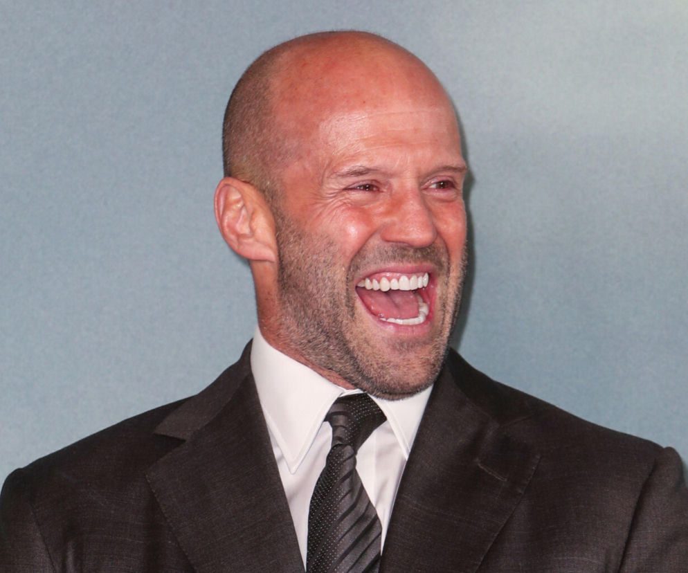 Jason Statham z uśmiechem na twarzy, ubrany w elegancki, ciemny garnitur i krawat. Aktor prezentuje radosne oblicze, co może zaskoczyć fanów jego typowych ról akcji. Tego typu informacje o gwiazdach i ich filmach znajdziesz na Super Seriale.