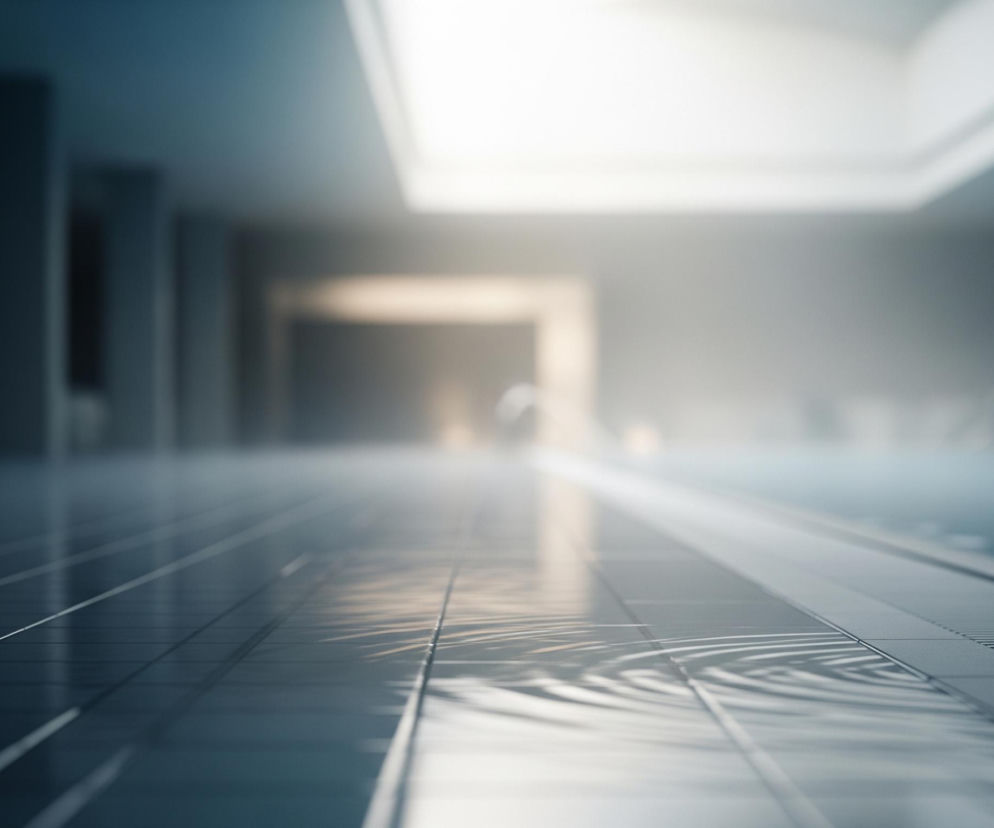 Ciemnoszara podłoga w pomieszczeniu z basenem zajmuje większą część kadru. Na jej powierzchni widoczne są płytki ułożone w regularną siatkę, a w lewej dolnej części podłogi odbijają się falujące refleksy świetlne z sufitu. Po prawej stronie, wzdłuż podłogi, ciągnie się ciemna, perforowana kratka odpływowa, za którą rozciąga się turkusowa tafla wody basenowej. W głębi pomieszczenia widoczne są niewyraźne, ciemne ściany i filary, a także jasne światło wpadające z dużego, prostokątnego świetlika w suficie.