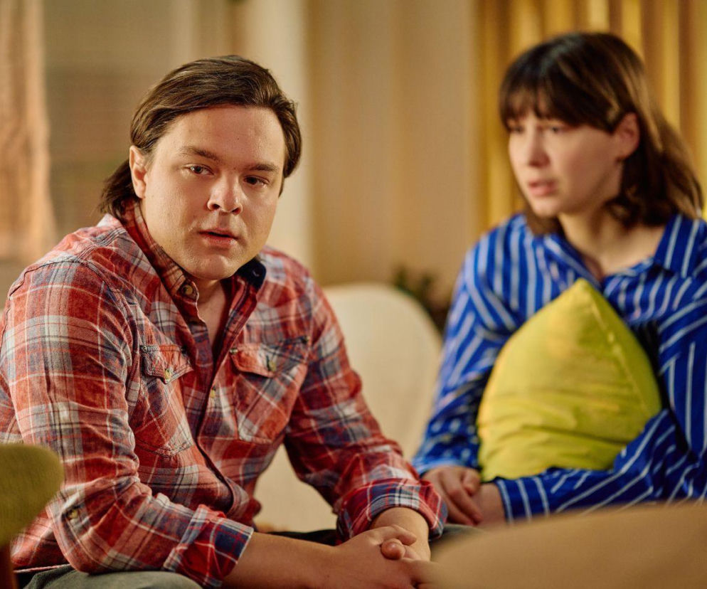 Młody mężczyzna w kraciastej koszuli patrzy wprost, siedząc obok kobiety trzymającej poduszkę. Scena sugeruje rozmowę lub problem, co odnosi się do informacji o zmianach w ramówce TVN i zniknięciu Na Wspólnej, o czym można przeczytać na Super Seriale.