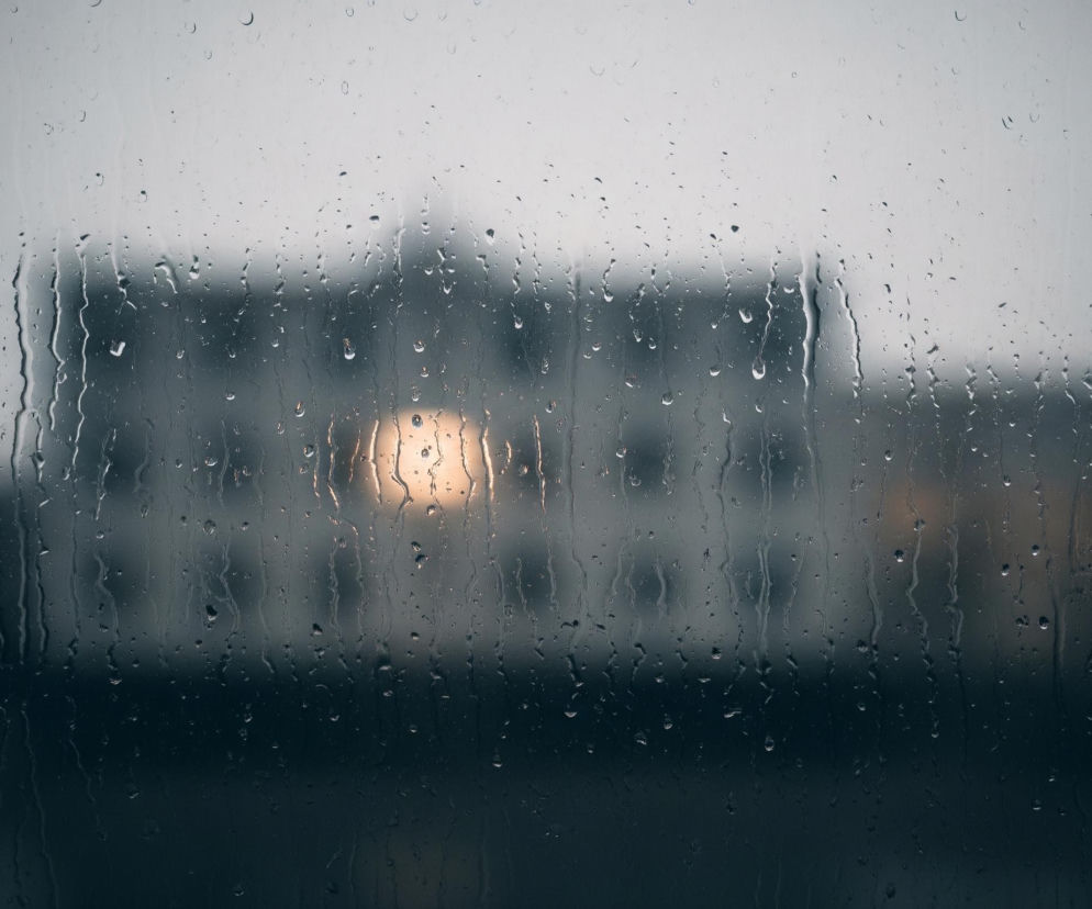 Przedni plan wypełnia szyba pokryta licznymi kroplami deszczu, które spływają w dół, tworząc pionowe smugi. W tle, za zamgloną szybą, widać rozmyty obraz budynku o jasnych ścianach, z którego okien wydobywa się ciepłe, żółtopomarańczowe światło. Górna część obrazu jest jaśniejsza, przyjmując odcienie szarości, podczas gdy dolna staje się ciemniejsza, przechodząc w granat.