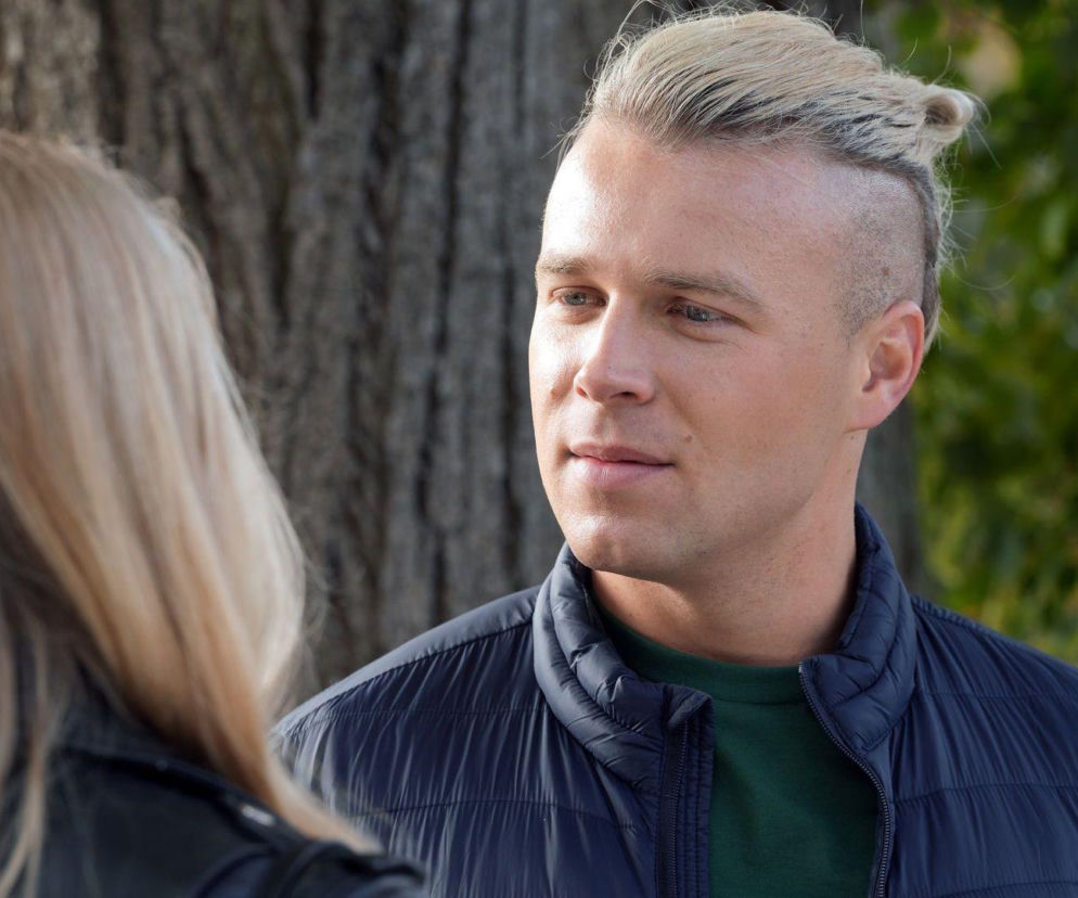 Patryk Jezierski (Konrad Skolimowski) w ciemnej kurtce i zielonej bluzie patrzy w lewo na blond kobietę, której tył głowy i włosy są widoczne na pierwszym planie. Patryk zerwie z Grażyną, bo nie wyzna mu miłości. Szczegóły serialu Barwy szczęścia znajdziesz na Super Seriale.