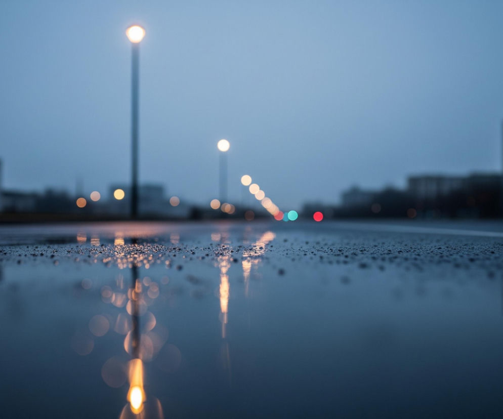 Mokra, ciemna nawierzchnia zajmuje dolną połowę obrazu, pokryta drobnymi kroplami wody w pierwszym planie i odbijająca światła uliczne oraz budynki w tle. Odblaski żółtych świateł latarni ulicznych, ułożonych w rzędzie w oddali, są widoczne na mokrej powierzchni. W tle, po obu stronach drogi, widać rozmyte kontury budynków. Po prawej stronie widać światła sygnalizacji świetlnej – zielone i czerwone.