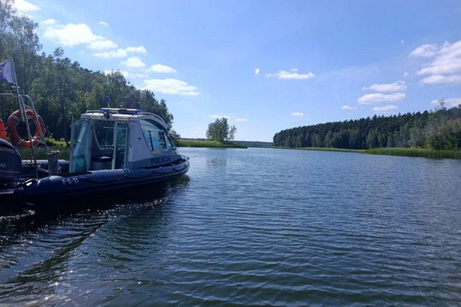Policyjna łódź motorowa na tle jeziora i zalesionego brzegu, z widocznym napisem „POLICJA”. Sezon wodny rozpoczęty, a policjanci będą dbać o bezpieczeństwo na wodzie. Więcej informacji o zasadach bezpieczeństwa przeczytasz na naszym portalu.