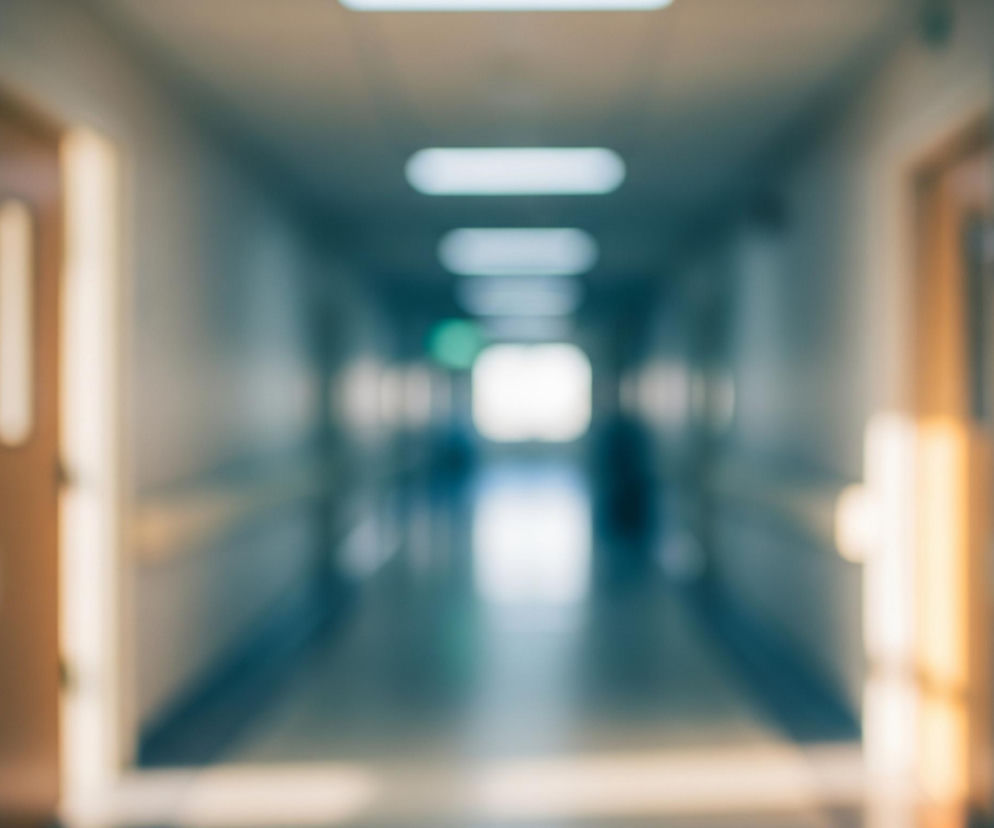 Rozmyty obraz przedstawia długi korytarz widziany z perspektywy pierwszej osoby, charakteryzujący się symetrycznym układem. Po obu stronach widoczne są jasnobrązowe drzwi z ciemnymi, prostokątnymi tabliczkami, a poniżej nich ciągną się poziome, beżowe barierki ochronne. Na suficie widać rzędy prostokątnych, jasno świecących lamp, które prowadzą wzrok w głąb pomieszczenia, gdzie intensywne białe światło sugeruje wyjście lub okno. Cała scena utrzymana jest w chłodnej tonacji błękitu i szarości, z ciepłymi akcentami brązu i beżu.