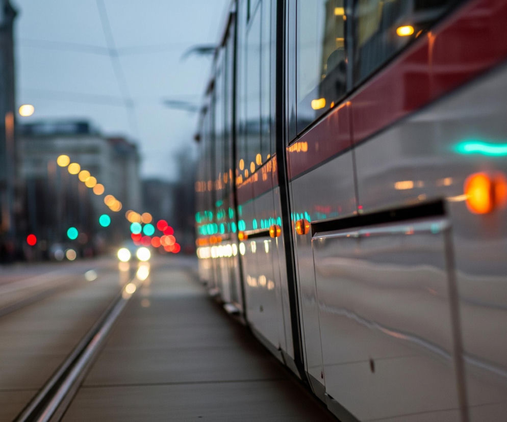 Biały tramwaj z czerwonym pasem pośrodku zajmuje prawą stronę kadru, z wyraźnymi oknami i odbijającymi światłami lamp, zarówno z wnętrza, jak i zewnętrznymi, pomarańczowymi i turkusowymi. Obok tramwaju biegną tory, które rozciągają się w lewą stronę zdjęcia, w głąb perspektywy. W tle, na końcu ulicy, znajdują się rozmyte budynki miejskie oraz liczne, zamazane światła uliczne i samochodowe w kolorach pomarańczowym, czerwonym i turkusowym, sugerujące wieczorną lub nocną scenerię.