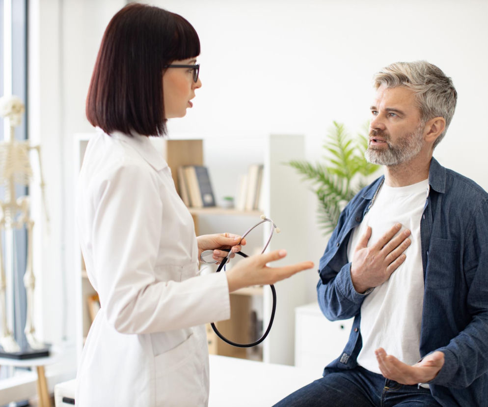 Kobieta lekarz w okularach i białym kitlu z stetoskopem w dłoni rozmawia z pacjentem, który trzyma rękę na klatce piersiowej, sygnalizując ból w okolicy serca. O tym, jak dbać o serce po zawale, przeczytasz na portalu Poradnik Zdrowie.