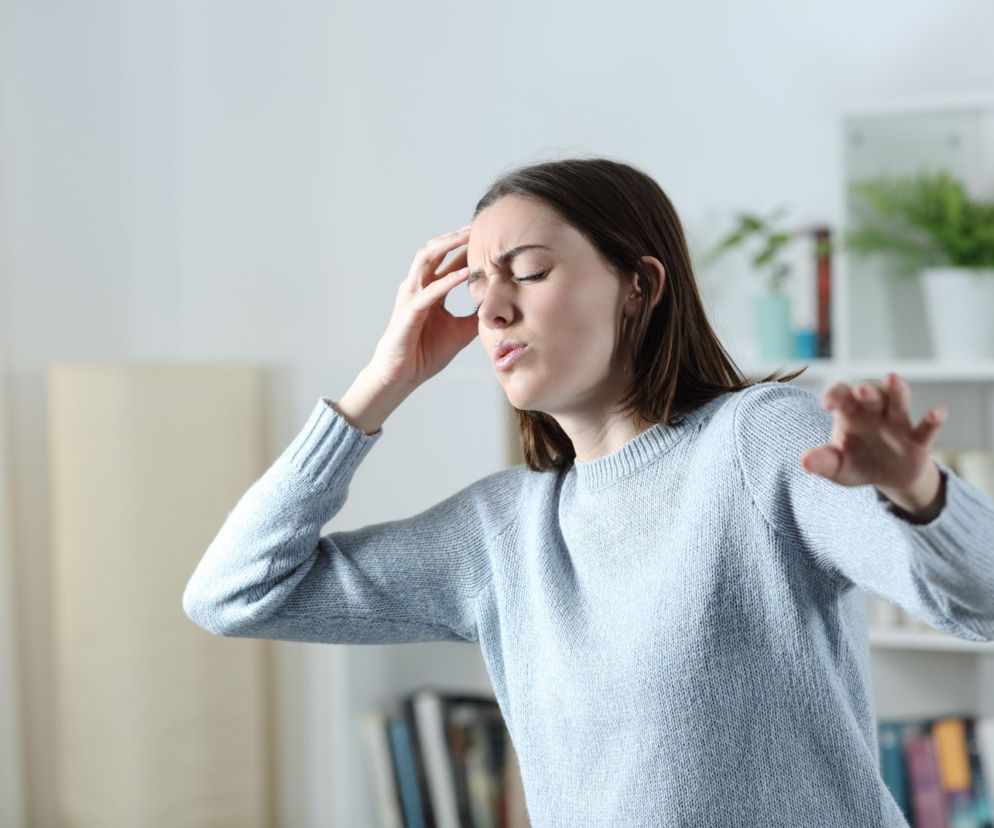 Młoda kobieta w jasnym swetrze łapie się za głowę, z wyraźnym grymasem bólu i zmęczenia, odczuwając zawroty głowy lub mroczki po szybkim wstaniu. Ilustruje problem hipotonii ortostatycznej, o której można przeczytać na portalu Poradnik Zdrowie.