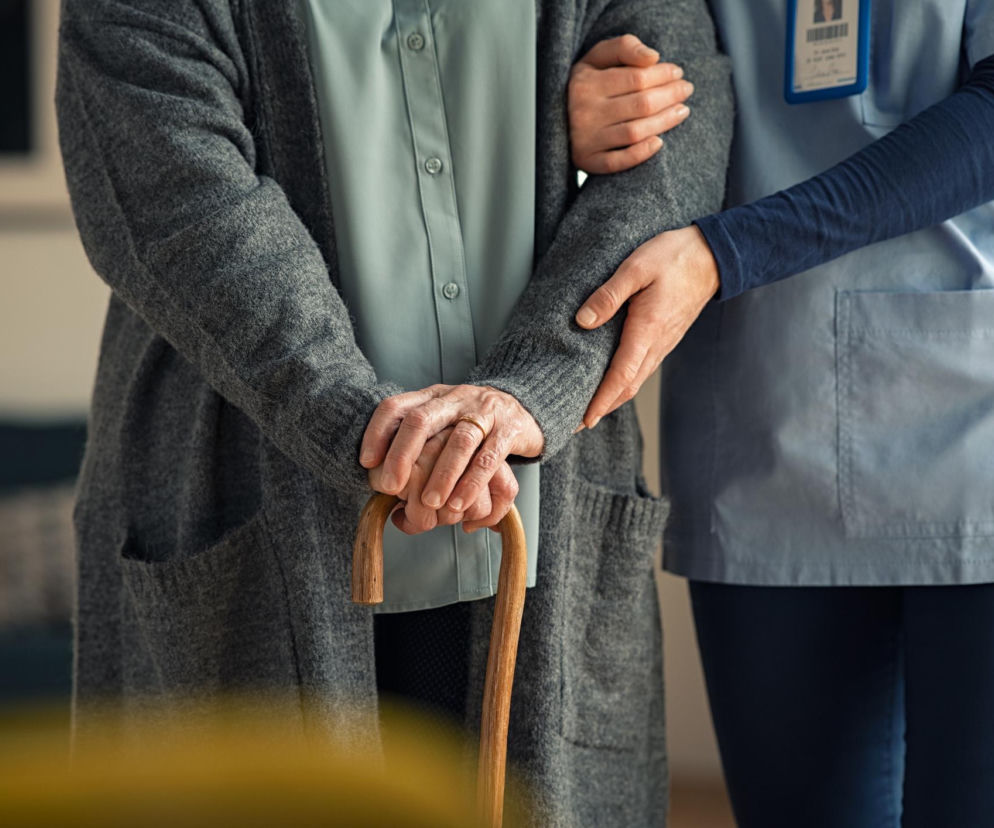 Opiekunka, w niebieskim uniformie, wspiera starszą kobietę, która opiera się na lasce, symbolizując potrzebę wsparcia w opiece nad seniorami. Temat braku rąk do pracy w sektorze opieki jest szeroko omawiany na Poradnik Zdrowie.