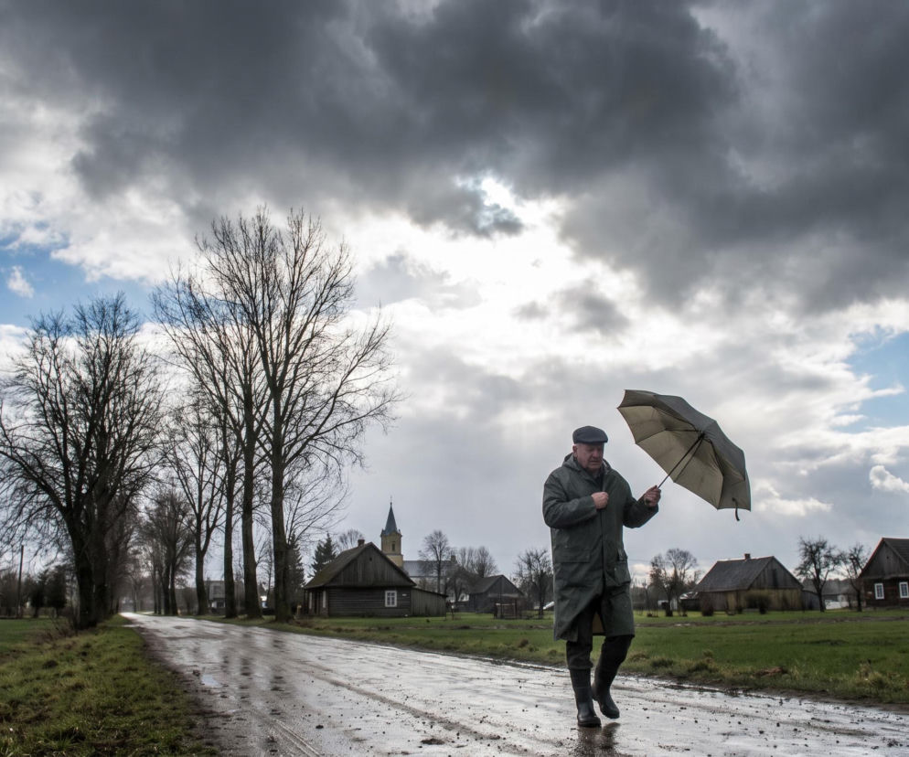 Mężczyzna w płaszczu z parasolem na mokrej drodze we wsi. O prognozie pogody i burzach przeczytasz na Eska.