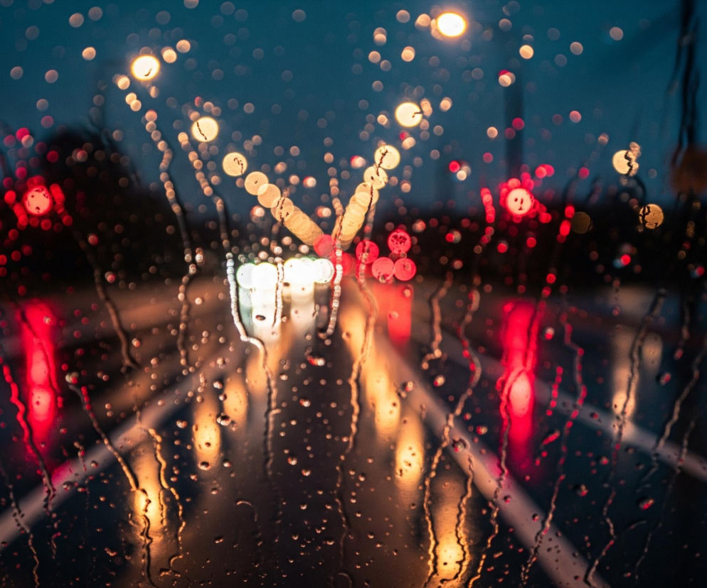 Widok na drogę podczas deszczowej nocy z perspektywy wnętrza samochodu, przez przednią szybę pokrytą licznymi kroplami wody. Światła miejskie i samochodowe są rozmyte, tworząc efekt bokeh w odcieniach czerwieni, pomarańczy i bieli. Główną część kadru zajmuje ciemna jezdnia, z rozmywającymi się białymi liniami pasów ruchu.