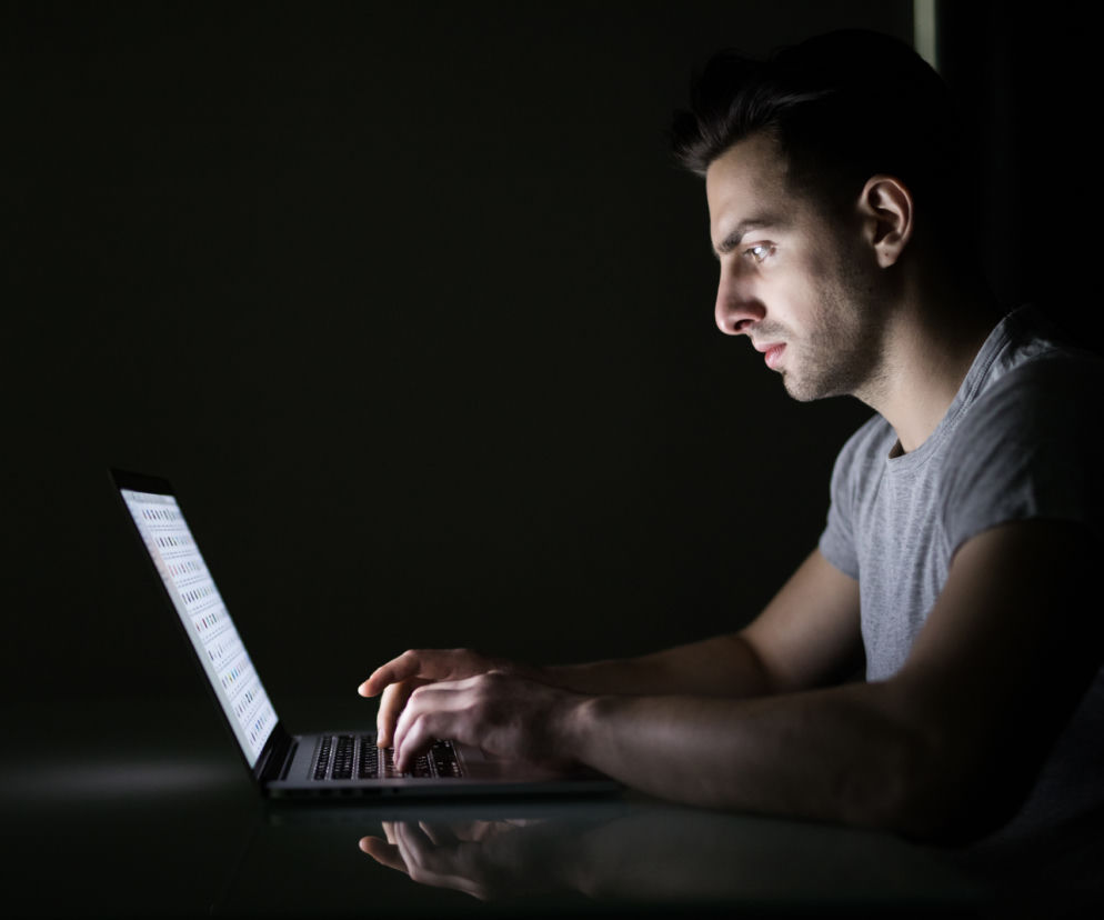 Mężczyzna pracuje nocą przy laptopie, skupiony na ekranie, w ciemnym otoczeniu, co symbolizuje ryzyko zaburzeń rytmu dobowego i ich wpływu na zdrowie. Na Poradnik Zdrowie znajdziesz informacje o skutkach pracy zmianowej.