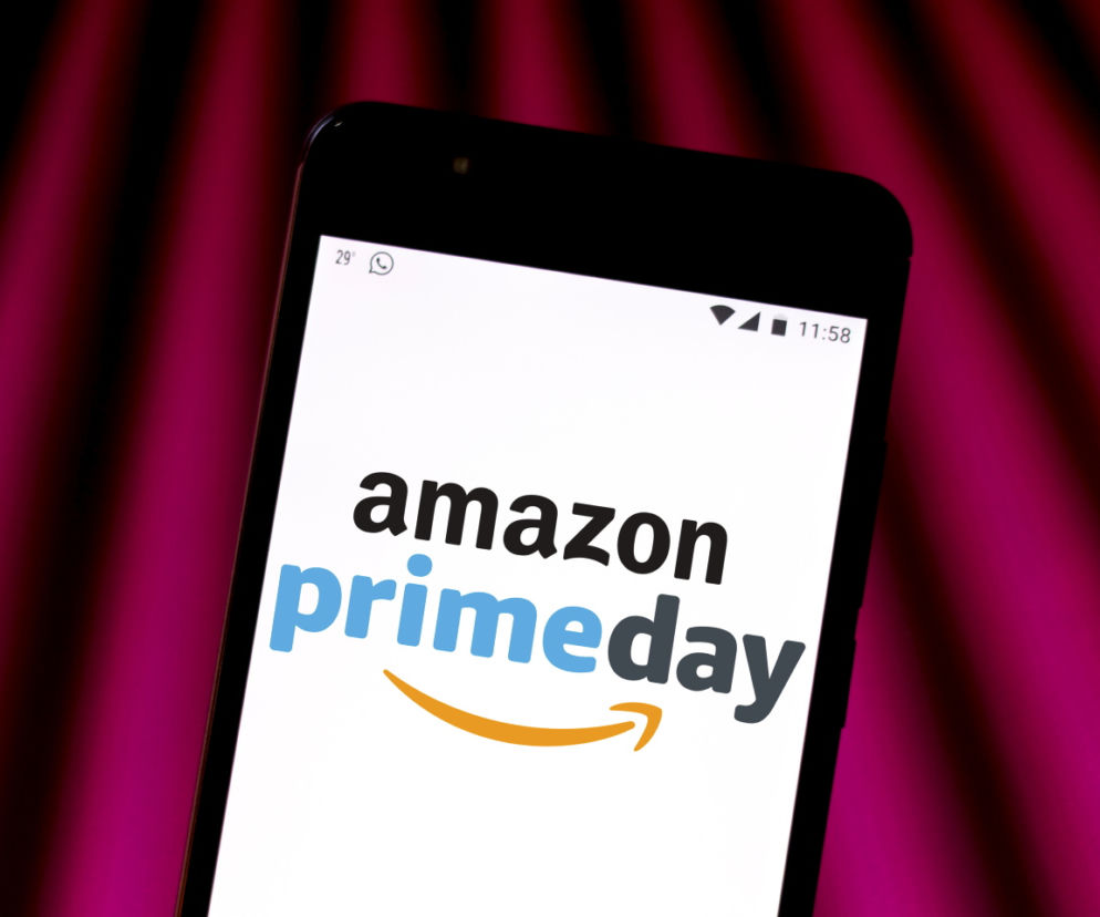 Smartfon wyświetlający logo amazon primeday na ekranie, symbolizujący zbliżające się wydarzenie zakupowe Amazon Prime Day, o którym przeczytasz na Super Biznes. Białe tło ekranu kontrastuje z ciemną obudową telefonu i rozmytym, fioletowo-różowym tłem.