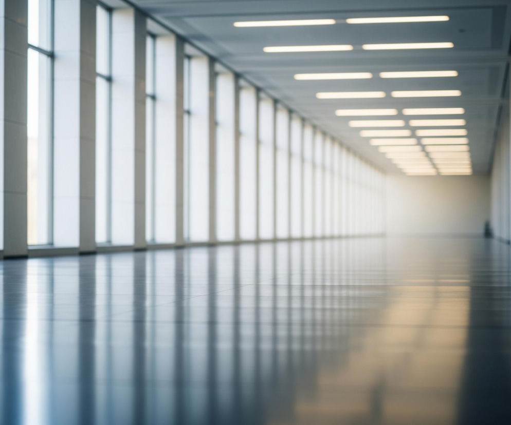 Długi, przestronny korytarz o nowoczesnej architekturze rozciąga się w głąb obrazu, znikając w jasnej, rozmytej bieli na horyzoncie. Po lewej stronie ciągnie się rząd wysokich okien z pionowymi, szarymi ramami, przez które wpada dzienne światło. Po prawej stronie znajdują się solidne, prostokątne filary, tworzące rytmiczną sekwencję. Na suficie widoczne są równoległe rzędy prostokątnych, świecących lamp, które również zbiegają się w oddali, potęgując wrażenie głębi. Podłoga wykonana jest z ciemnego, lśniącego materiału, który doskonale odbija światło z okien i lamp, tworząc długie, pionowe refleksy. Całość utrzymana jest w chłodnych odcieniach szarości, bieli i błękitu, z delikatnymi ciepłymi akcentami światła.