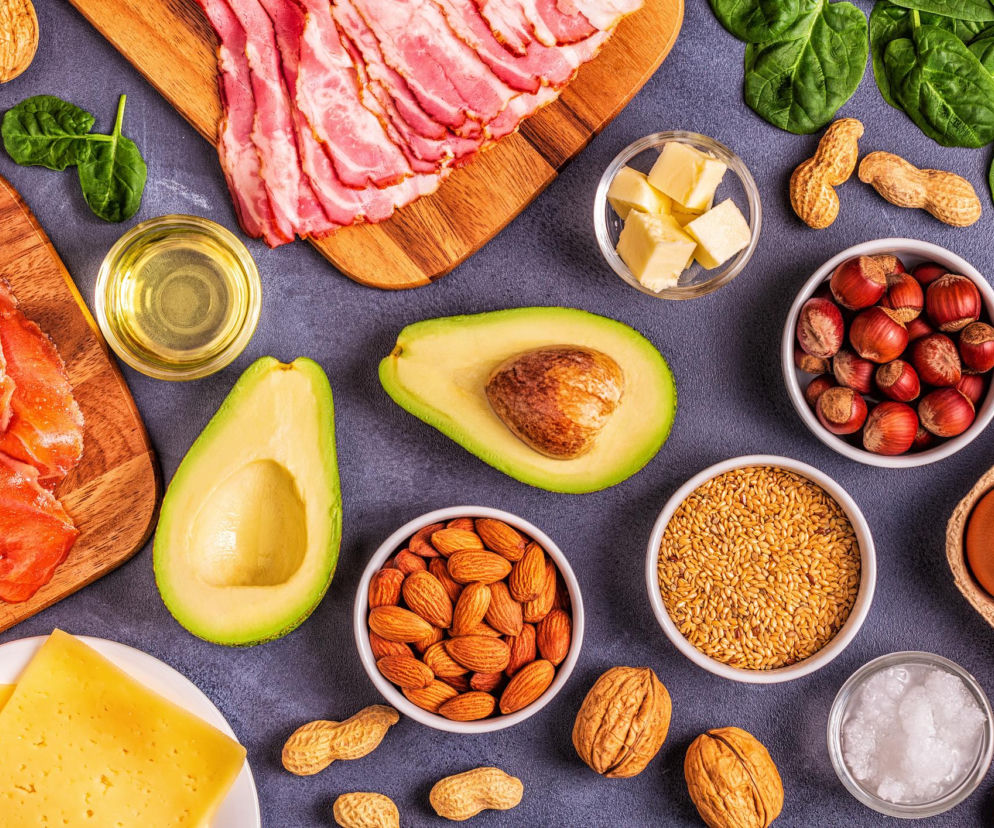 Kompozycja różnych zdrowych tłuszczów na ciemnym tle, w tym awokado, łosoś, boczek, ser, orzechy, nasiona i jajka. Zdjęcie ilustruje różnorodność produktów bogatych w tłuszcze, które są korzystne dla zdrowia, o czym przeczytasz więcej na Poradnik Zdrowie.